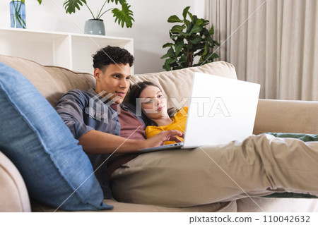 Happy diverse couple lying on couch and using laptop in living room at home, copy space 110042632