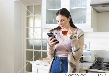 Focused caucasian woman standing, holding tea cup, using smartphone in kitchen at home, copy space 110042634