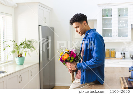 Happy biracial man holding flowers in kitchen at home, copy space 110042690