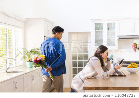 Diverse couple holding flowers behind back and using smartphone in kitchen at home, copy space Diverse couple holding flowers behind back and using smartphone in kitchen at home, copy space 110042722
