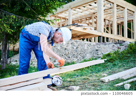 Carpenter constructing wooden-framed house. Man measures distances using tape measure while dressed in work attire and helmet. The idea behind ecologically sound, contemporary construction. 110046605