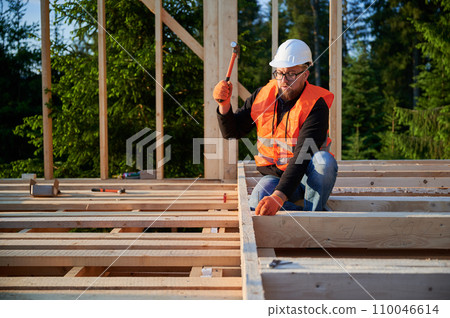 Carpenter in process of building wooden-framed dwelling adjacent to woods. Bespectacled and bearded man pounds in nails with hammer. Concept of contemporary ecological building. 110046614