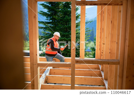 Carpenter constructing two-story wooden frame house near forest. Man hammering nails into the structure, wearing protective helmet and construction vest. Concept of modern ecological construction. Carpenter constructing two-story wooden frame house near forest. Man hammering nails into the structure, wearing protective helmet and construction vest. Concept of modern ecological construction. 110046617
