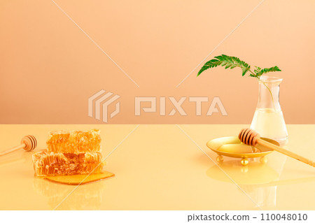 Natural raw beeswax is stacked on top of each other, a fern leaf is inserted into an erlenmeyer flask, a wooden honey spoon is placed next to a gold plate on a pastel background. 110048010