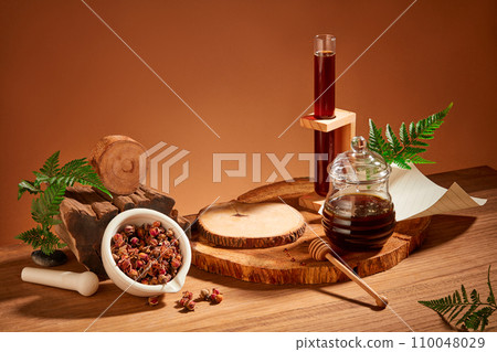 Honey is stored in glassware, dried rose buds in a mini ceramic mortar, fern leaves and logs are placed on a wooden table. Brown background for advertising. Honey is stored in glassware, dried rose buds in a mini ceramic mortar, fern leaves and logs are placed on a wooden table. Brown background for advertising. 110048029