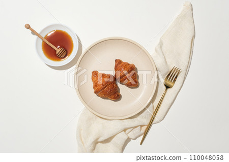 Two croissants are displayed on round ceramic dish, a metal fork, honey and a honey drizzle on a white plate and a cloth towel on a white background. Advertising with natural ingredients. Two croissants are displayed on round ceramic dish, a metal fork, honey and a honey drizzle on a white plate and a cloth towel on a white background. Advertising with natural ingredients. 110048058