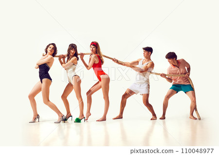 Game on. Young cheerful people, friends in swimsuits playing on beach, pulling rope against white background. Fun and joy Game on. Young cheerful people, friends in swimsuits playing on beach, pulling rope against white background. Fun and joy 110048977