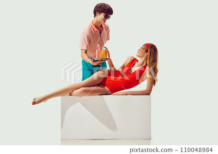Summer seaside resort. Young man serving drink to beautiful woman in swimsuit sunbathing against white background 110048984