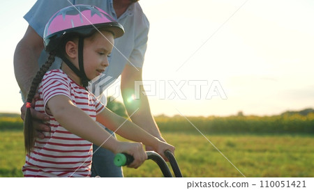Concentrated little girl rides bicycle caring father holds vehicle in field 110051421