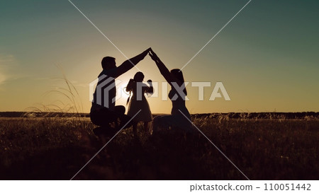 Silhouettes of parents intertwine above daughter head in meadow at sunset twilight 110051442