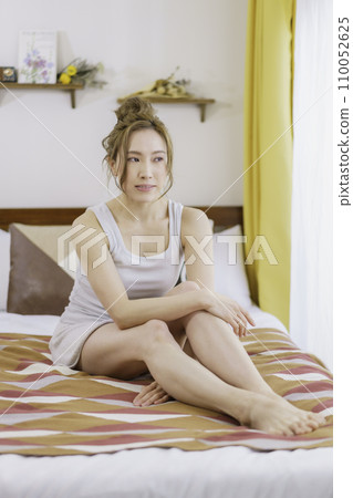 Portrait of a brown-haired young woman sitting on bed, vertical position Portrait of a brown-haired young woman sitting on bed, vertical position 110052625