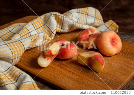 Cutting board with halves and three whole saturn peaches or flat peaches on wooden background with yellow kitchen towel.. 110052933