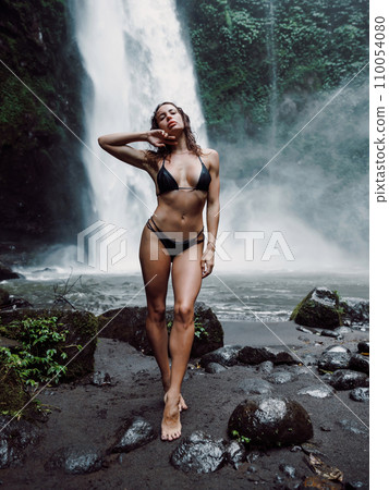 Slim woman in bikini staying near waterfall in Bali. Traveler girl posing on river with powerfull waterfall Slim woman in bikini staying near waterfall in Bali. Traveler girl posing on river with powerfull waterfall 110054080