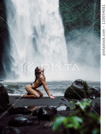 Woman in swimsuit posing near powerful waterfall. Traveler girl on waterfall in Bali Woman in swimsuit posing near powerful waterfall. Traveler girl on waterfall in Bali 110054081
