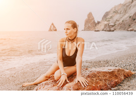 Woman travel portrait. Happy woman with long hair poses on a red volcanic rock at the beach. Close up portrait cute woman in black bikini, smiles at the camera, with the sea in the background. Woman travel portrait. Happy woman with long hair poses on a red volcanic rock at the beach. Close up portrait cute woman in black bikini, smiles at the camera, with the sea in the background. 110056439