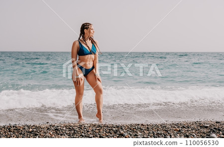 A tourist woman in blue bikini on beach enjoys the turquoise sea during her summer holiday. Rear view. Beach vacation. woman with braids dreadlocks standing with her arms raised enjoying beach ocean. 110056530