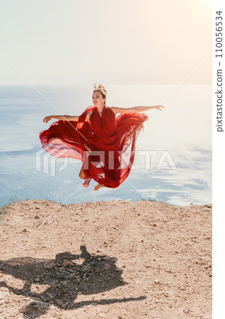 Woman in red dress on sea. Side view a Young beautiful sensual woman in a red long dress posing on a rock high above the sea on sunset. Girl on the nature on blue sky background. Fashion photo. 110056534