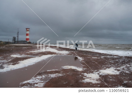 Lighthouse on the shore. Difficult way to the lighthouse. A man in motion, seen from the back, is walking with difficulty along the shore of a stormy sea. 110056921