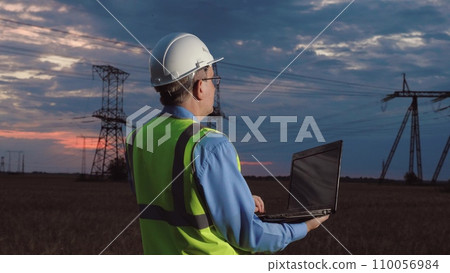 Electrician with helmet checkup powerlines network with laptop in evening field. Skilled engineer does diagnostic tests of power generation substation with laptop in sunset country field 110056984