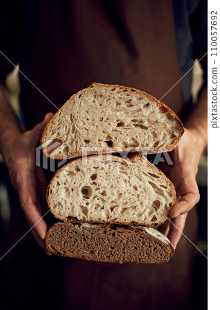Bakery man, chef in brown apron holds in hands two halves of just whole grain hot loaf of bread with golden brown crispy crust. Concept of bakery shop 110057692