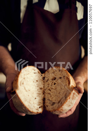 Man in brown apron holds in hands two halves of just prepared, delicious, hot homemade loaf of bread with golden brown crispy crust. Man in brown apron holds in hands two halves of just prepared, delicious, hot homemade loaf of bread with golden brown crispy crust. 110057697