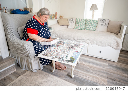 An elderly woman collecting puzzle while sitting in a chair in her bright room at home. 110057947