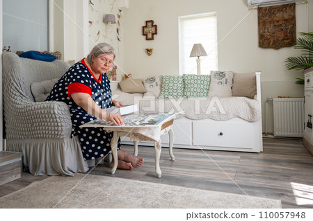 An elderly woman collecting puzzle while sitting in a chair in her bright room at home. 110057948