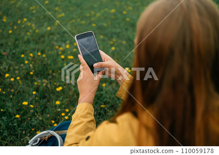 Back view of young woman touching cell phone and sits on meadow 110059178
