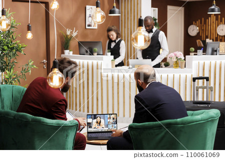 Business partners on videocall at hotel, talking to colleagues on online teleconference meeting. People in suits travelling for work and using videoconference call on laptop, lobby area. Business partners on videocall at hotel, talking to colleagues on online teleconference meeting. People in suits travelling for work and using videoconference call on laptop, lobby area. 110061069
