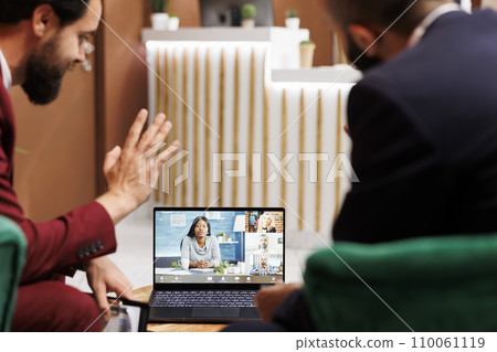 Two businessmen meet on videocall with other international company representatives, using laptop in hotel lobby. Management team talking on online telework conference, executive board. Two businessmen meet on videocall with other international company representatives, using laptop in hotel lobby. Management team talking on online telework conference, executive board. 110061119
