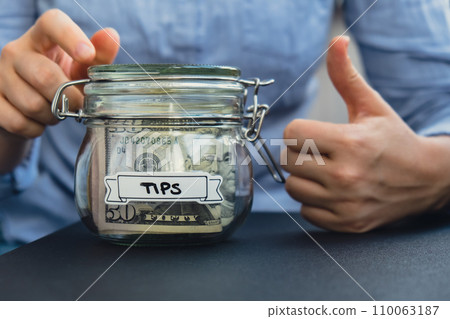 Unrecognizable woman holding Glass jar full of American currency dollars cash banknote with text TIPS. Preparation saving money. Moderate consumption and economy 110063187