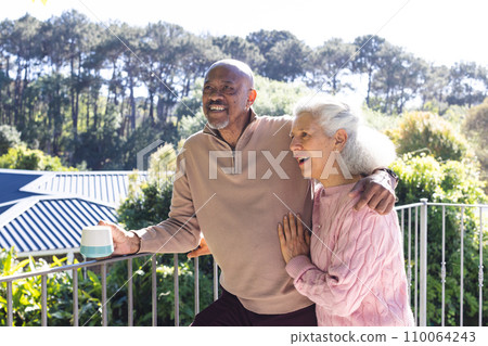 Happy diverse senior couple holding cup of coffee and embracing on sunny terrace Happy diverse senior couple holding cup of coffee and embracing on sunny terrace 110064243