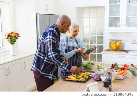 Diverse senior couple preparing meal using tablet in kitchen Diverse senior couple preparing meal using tablet in kitchen 110064342