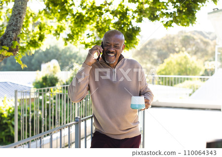 Happy african american senior man holding cup of coffee and talking on smartphone on sunny terrace 110064343
