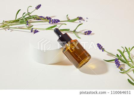 A bottle of serum is leaning against a white podium, surrounded by lavender flowers on a white background. Lavender essential oil fades dark spots and helps even out skin tone. 110064651