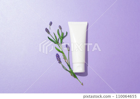 A tulip branch is placed next to an unlabeled cosmetic tube on a pastel purple background. Natural cosmetics concept with lavender extract. Exquisite space. A tulip branch is placed next to an unlabeled cosmetic tube on a pastel purple background. Natural cosmetics concept with lavender extract. Exquisite space. 110064655