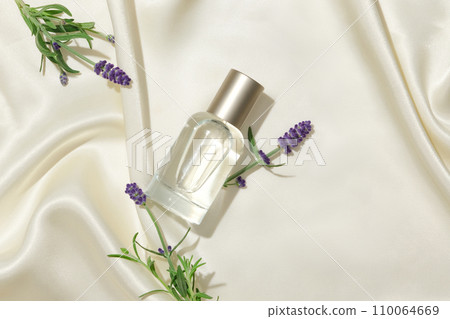 A bottle of unbranded perfume and fresh lavender flowers are placed on a silk background. The purple color stands out on the white background. Exquisite cosmetic mockup. 110064669
