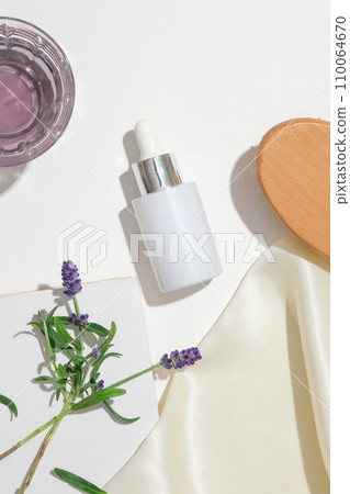 A bottle of serum without label, a glass containing light purple liquid and props on a white background. The sophisticated space is suitable for advertising natural cosmetics. 110064670