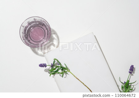 A glass containing a transparent light purple liquid, a white podium and fresh lavender flowers displayed on a minimalist white background. Space for advertising. 110064672
