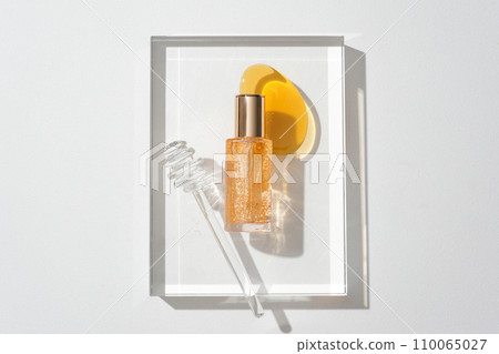 A serum bottle with yellow essence particles, honey drops and a honey drizzle on a glass podium on a white background. Cosmetic mockup for advertising. 110065027