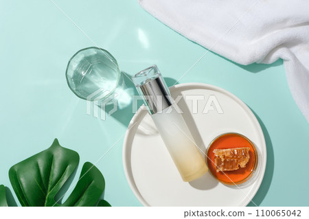 Honey and an unlabeled cosmetic bottle are displayed on a white ceramic plate, a glass of water, green leaves and a towel on a blue background. View from above. Honey and an unlabeled cosmetic bottle are displayed on a white ceramic plate, a glass of water, green leaves and a towel on a blue background. View from above. 110065042