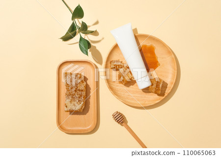 Beeswax is stored on a wooden tray, an unlabeled cosmetic tube and honey on a round wooden plate, a honey drizzle and green leaves on a pastel background. Ideal space for cosmetics display. 110065063