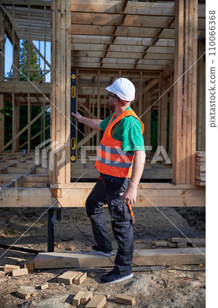 Carpenter constructing two-storey wooden frame house. Man inspects walls for levelness using spirit level, wearing protective overalls, helmet and vest. Concept of modern ecological construction. Carpenter constructing two-storey wooden frame house. Man inspects walls for levelness using spirit level, wearing protective overalls, helmet and vest. Concept of modern ecological construction. 110066368