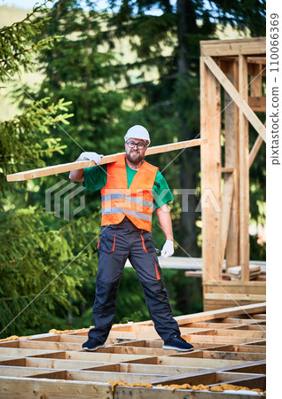 Carpenter building wooden frame house near the forest. Happy man holds large plank on his shoulder, dressed in work clothes and helmet. Concept of contemporary and sustainable construction. Carpenter building wooden frame house near the forest. Happy man holds large plank on his shoulder, dressed in work clothes and helmet. Concept of contemporary and sustainable construction. 110066369