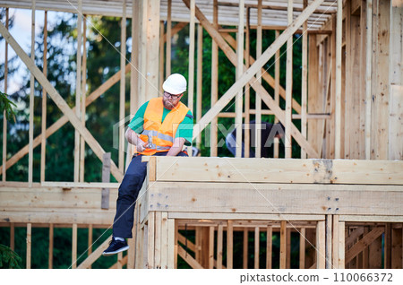 Carpenter constructing two-story wooden frame house near the forest. Bearded man in glasses hammering nails into structure while wearing protective helmet. Concept of modern ecological construction. Carpenter constructing two-story wooden frame house near the forest. Bearded man in glasses hammering nails into structure while wearing protective helmet. Concept of modern ecological construction. 110066372