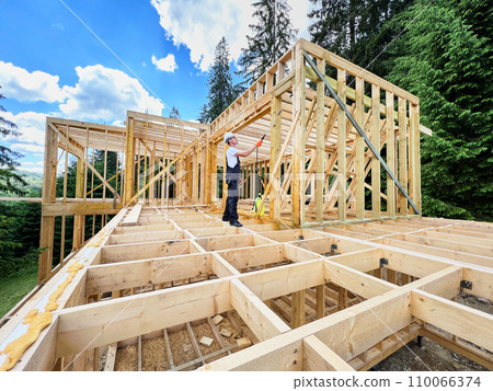 Laborer constructing wooden frame house near forest. Man treating woods, applying fire retardant using sprayer, while dressed in protective suit, helmet. Concept of modern eco-friendly construction. Laborer constructing wooden frame house near forest. Man treating woods, applying fire retardant using sprayer, while dressed in protective suit, helmet. Concept of modern eco-friendly construction. 110066374