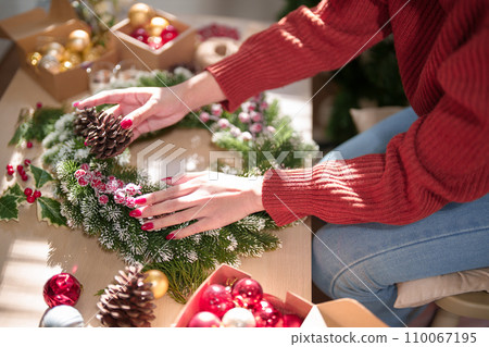Young asian woman in sweater making christmas wreath on wood table and decorations with pine cone and red berries while prepare ornaments to celebrate for christmas festive holiday and winter seasons 110067195
