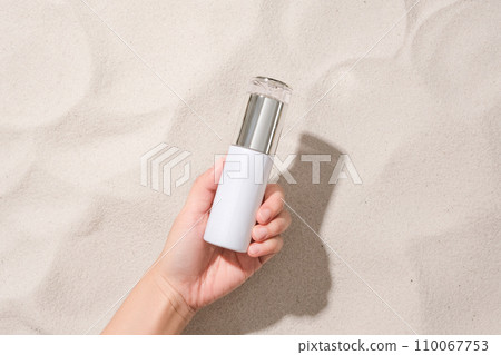 A hand is holding an unlabeled cosmetic bottle on the sandy background. Sophisticated space with a top-down view. Cosmetics advertising. 110067753