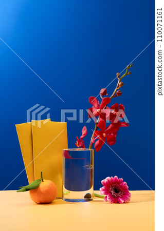 A fresh orchid is placed in a glass, two yellow envelopes, a gerbera flower and a tangerine are displayed on a beige table with a blue background. Copy space with front view. 110071161