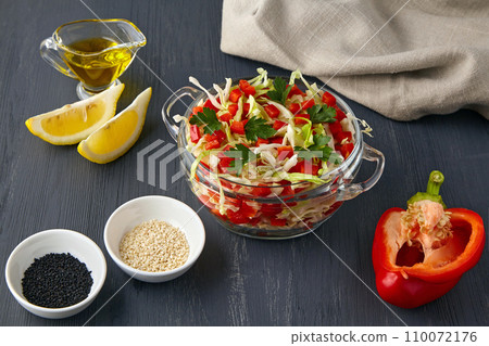 Fresh vegetable salad of cabbage and sweet peppers in a glass bowl on a black wooden background surrounded by ingredients 110072176